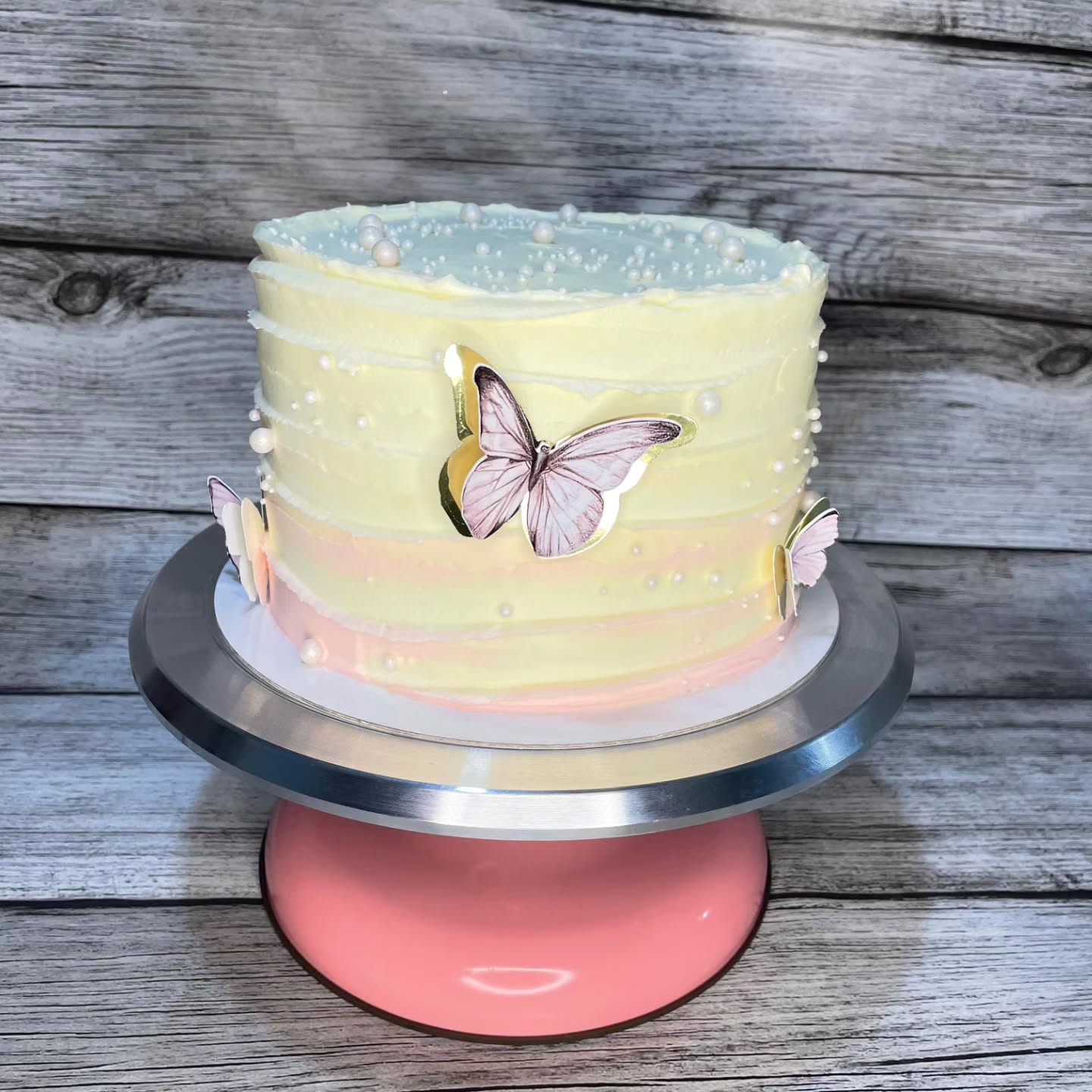 Pastel decorado con glaseado en tonos amarillo y rosa, adornado con mariposas de papel y perlas comestibles, presentado en un pedestal rosa. Ideal para celebraciones y eventos especiales.